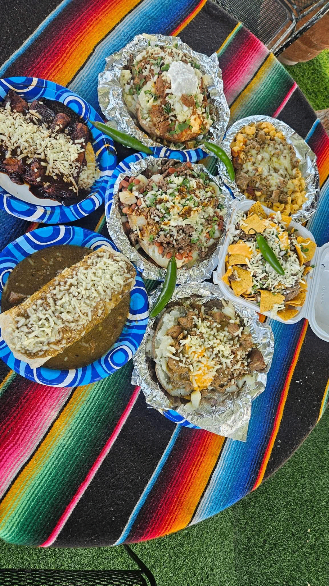 Full table spread of wet burritos, loaded baked potatoes, super nachos, mac & cheese chili verde and enchiladas on a striped Mexican blanket